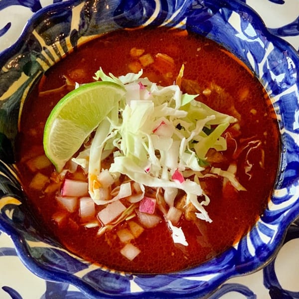 A vibrant red Mexican soup, garnished with shredded cabbage, diced radishes, and a lime wedge.