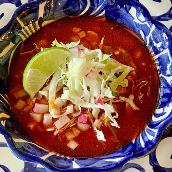 A vibrant red bowl of Favela's Mexican Grill's pozole, garnished with shredded cabbage, diced radish, and a lime wedge.