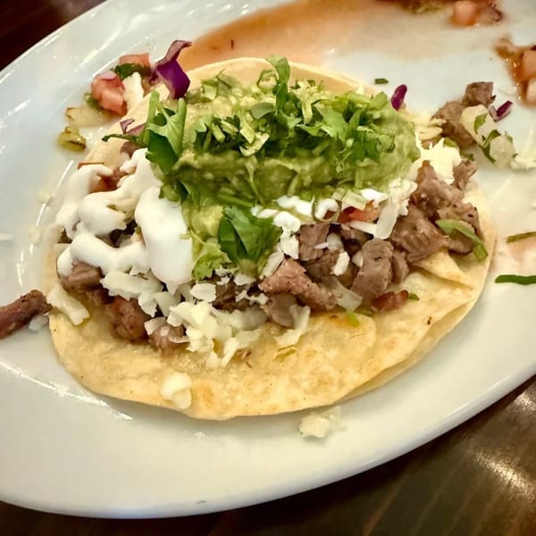 A Favela's Mexican Grill carne asada taco with guacamole, sour cream, and cheese on a white plate.