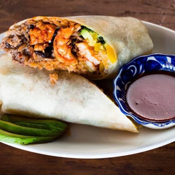 A large Favela's Mexican Grill burrito, cut in half, revealing shrimp, rice, and vegetables, with a side of sauce.
