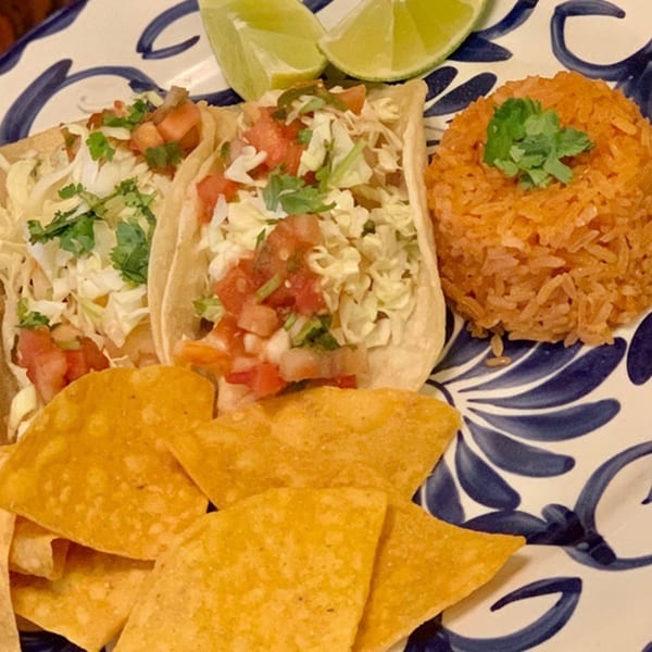 Two Favela's Mexican Grill tacos with rice, chips, and lime wedges on a blue and white plate.