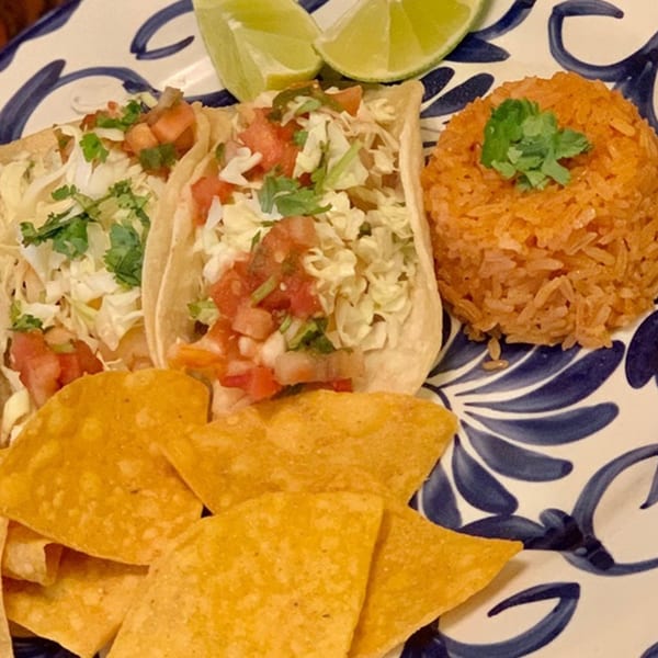 Two Favela's Mexican Grill tacos with rice, tortilla chips, and lime wedges on a blue and white plate.