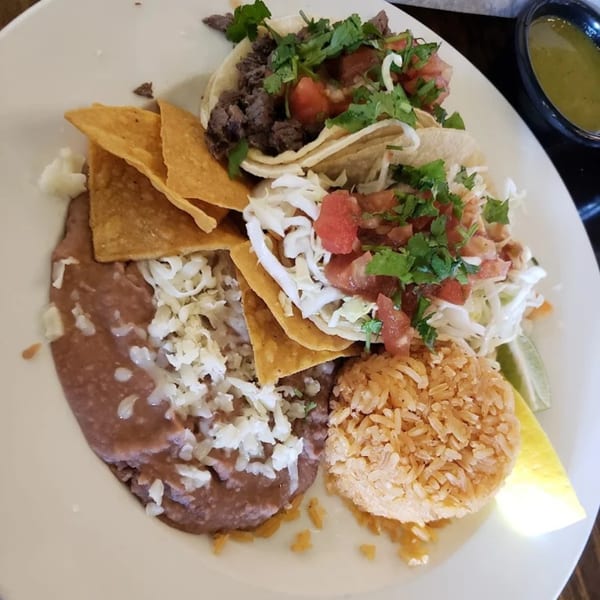 A plate of Favela's Mexican Grill tacos, refried beans, rice, tortilla chips, and a lime wedge.