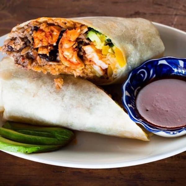 A Favela's Mexican Grill burrito, cut in half, revealing shrimp, rice, and avocado, with a side of sauce.